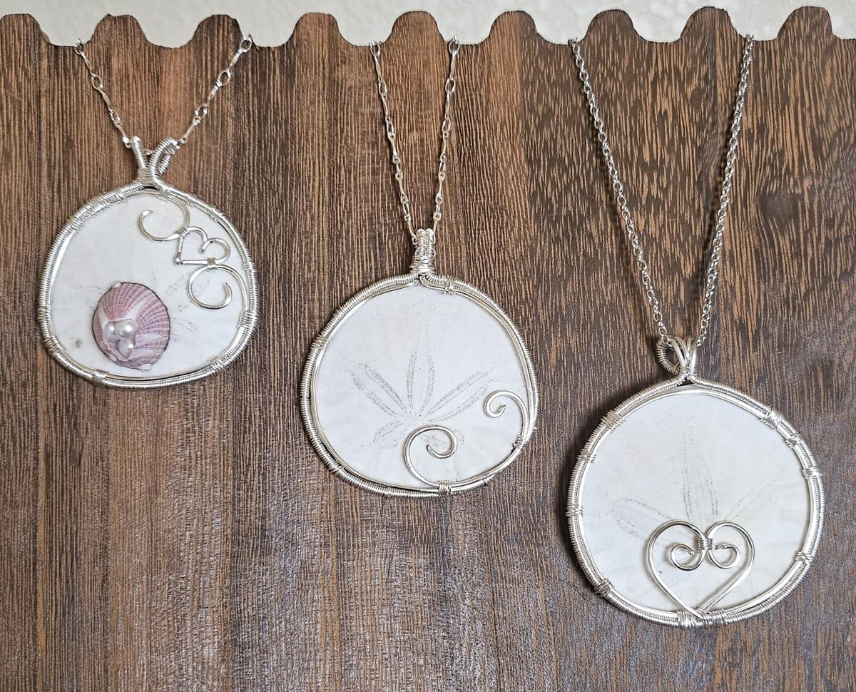 Three wire-wrapped pendants on a wooden board — a shell and two sand dollars, each caught in silver wire.