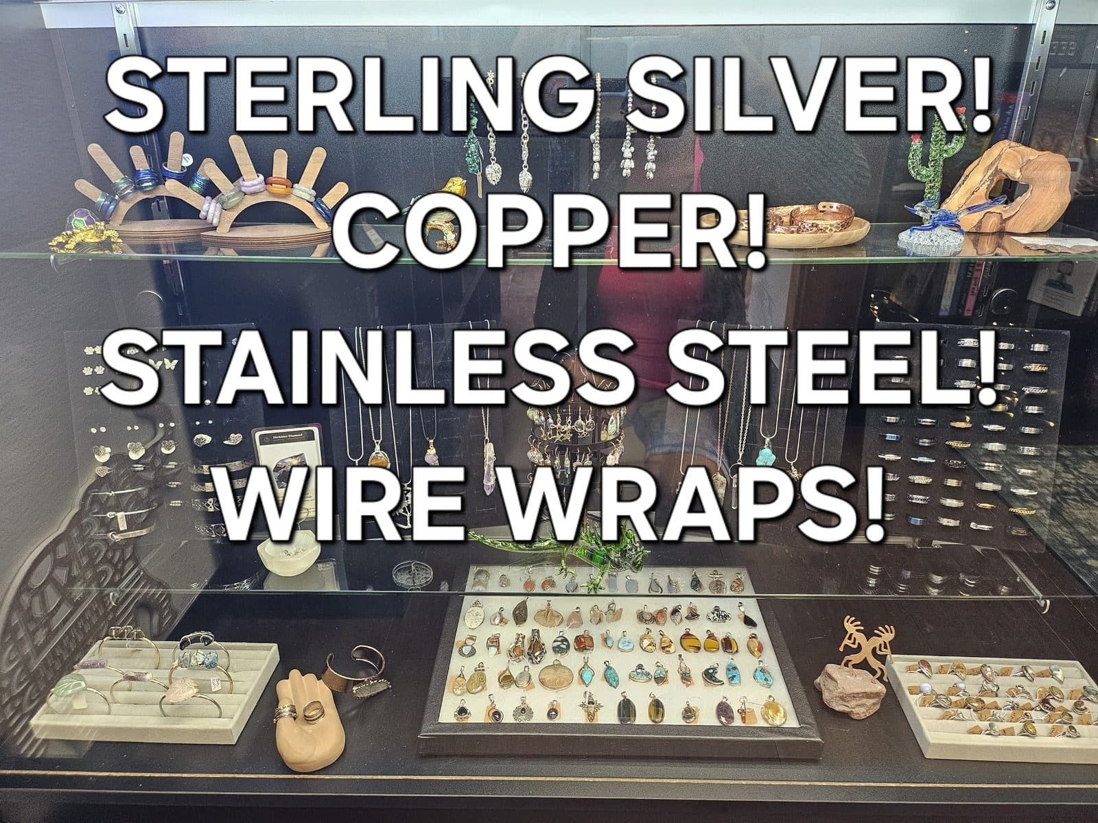 A lit glass display case of pendants, rings and wire-wrapped stones, captioned “Sterling silver! Copper! Stainless steel! Wire wraps!”.